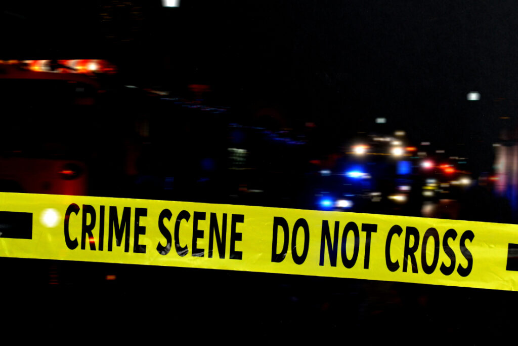Yellow police tape with the words 'CRIME SCENE DO NOT CROSS' in the foreground, with blurred emergency response vehicles and their flashing lights in the background during a night scene.