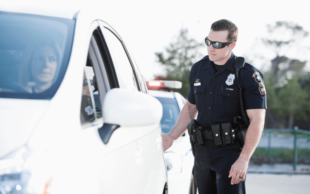 A police officer, wearing sunglasses and full uniform, approaches the driver's side of a white sedan during a routine traffic stop.