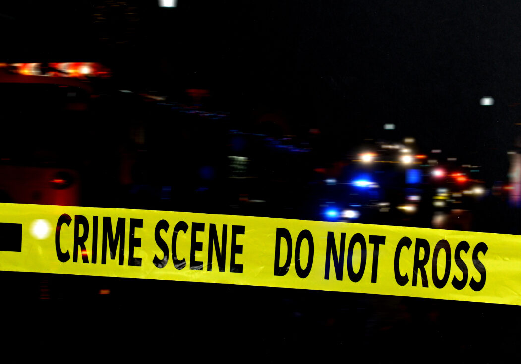 Yellow police tape with the words 'CRIME SCENE DO NOT CROSS' in the foreground, with blurred emergency response vehicles and their flashing lights in the background during a night scene.
