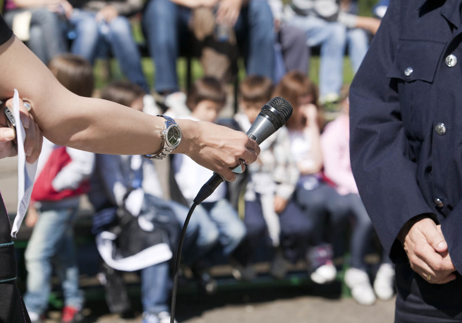 Cropped view of a woman's hand holding a microphone towards a person in police uniform, with a blurry group of seated children in the background.