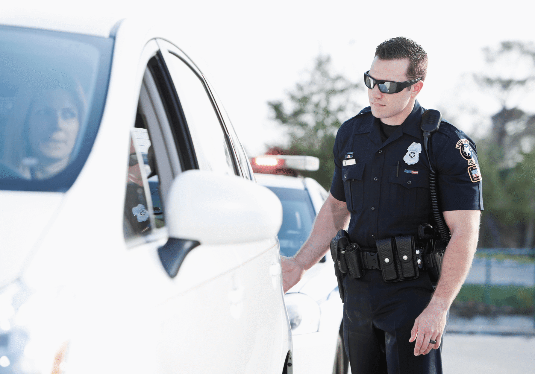A police officer, wearing sunglasses and full uniform, approaches the driver's side of a white sedan during a routine traffic stop.